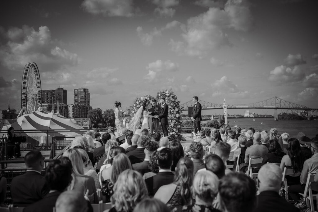 Black and white photograph of an outdoor wedding ceremony at Le Belvédère in the Old Port of Montreal, captured by wedding photographer Jeff Gagnon, featuring the couple exchanging vows under a floral arch with views of the Ferris wheel, Jacques Cartier Bridge, and the waterfront in the background.